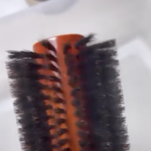 Close-up of a round hairbrush with dark bristles and a wooden handle, positioned against a white blurred background.
